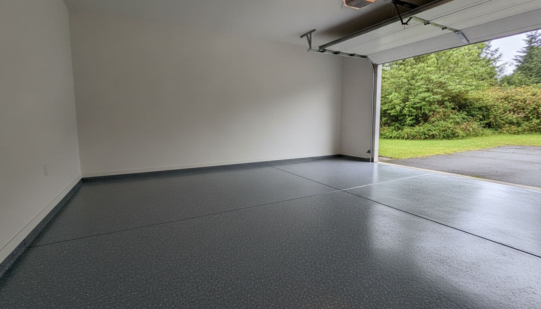 A charcoal grey coated garage floor with the door open onto a wet West Coast driveway.